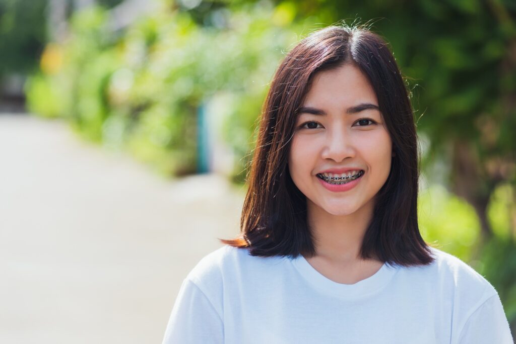 Young woman with long dark hair smiles to show her braces outdoors in a green park, for Ray + Swenson Orthodontics