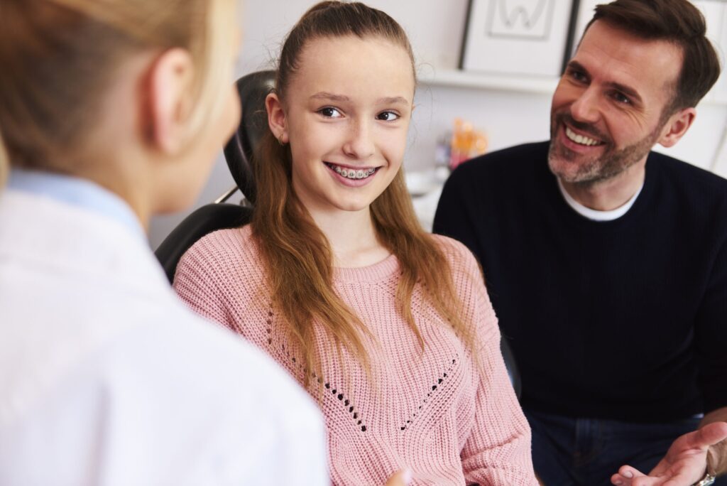 Smiling teen girl with braces sits in an exam chair beside her father during a consultation at Ray + Swenson Orthodontics