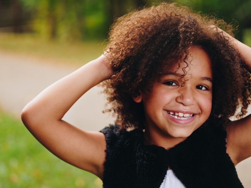 Young girl with curly hair smiles with her hand on her head, showing a healthy smile, for Ray + Swenson Orthodontics