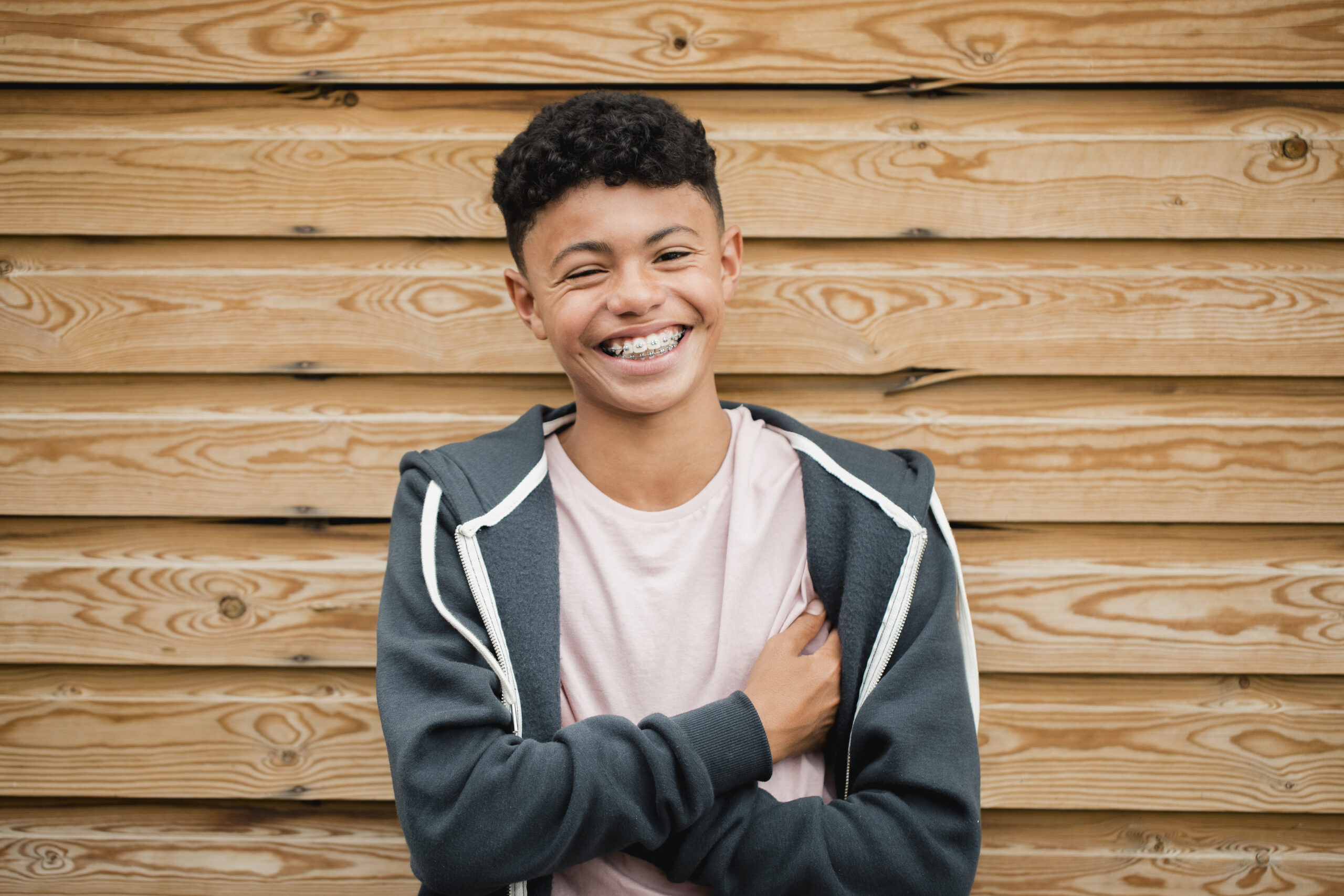 Teen boy in a gray hoodie smiles to show his braces in front of a wood wall, for Ray + Swenson Orthodontics